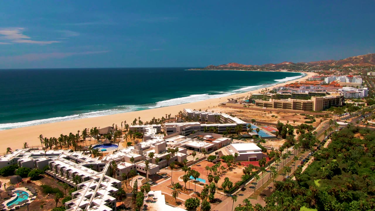 Aerial coastline view with resort buildings beside a long sandy beach and ocean surf.