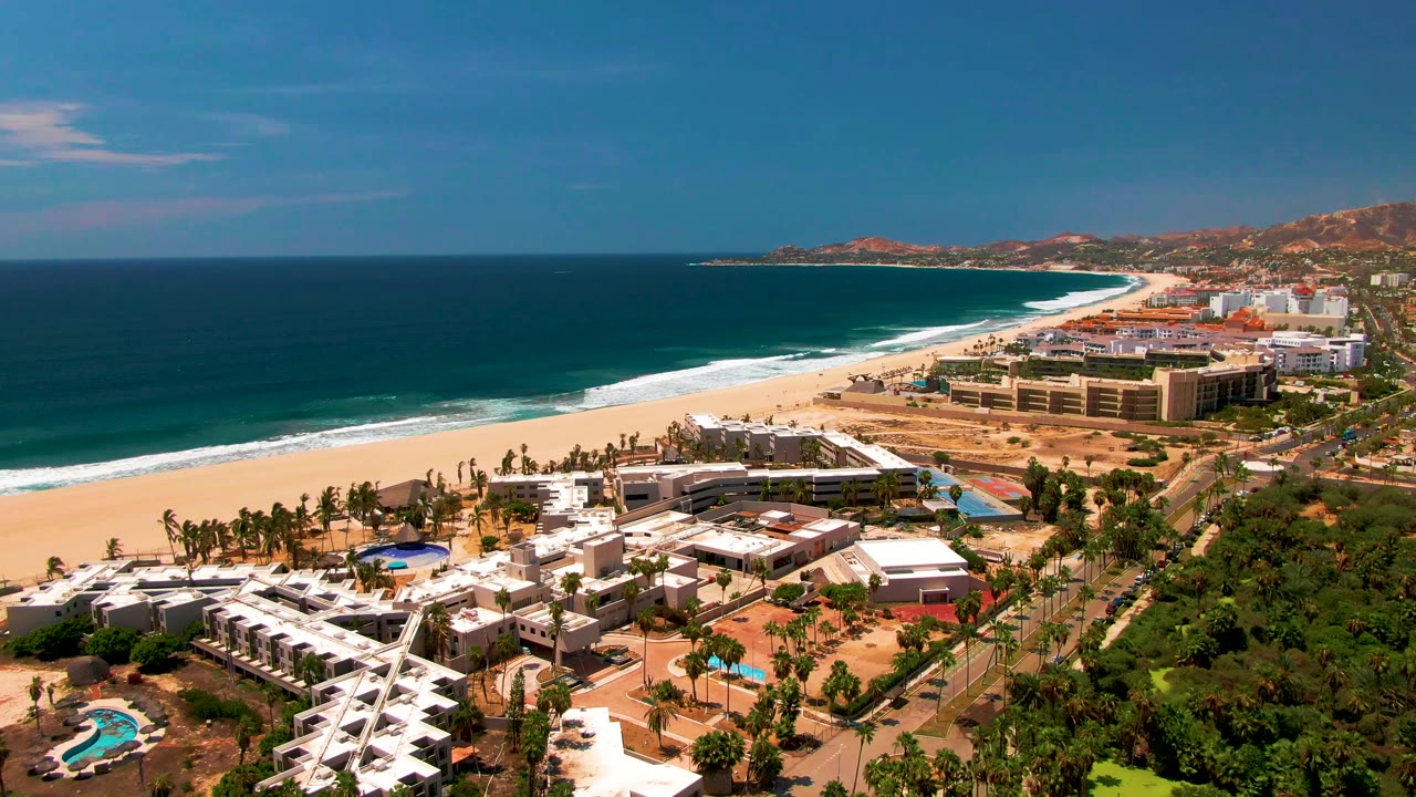 Elevated San Jose Del Cabo beach scene with ocean filling the left side, a strip of white surf, tan sand, large resort buildings, palms, green foreground trees, and hills behind town.