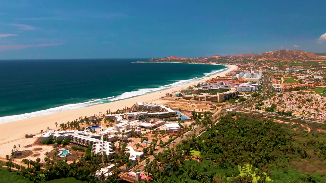 Aerial view over San Jose del Cabo with a wide sandy beach beside dark blue ocean, resort buildings, green vegetation, inland hills, and no visible people.