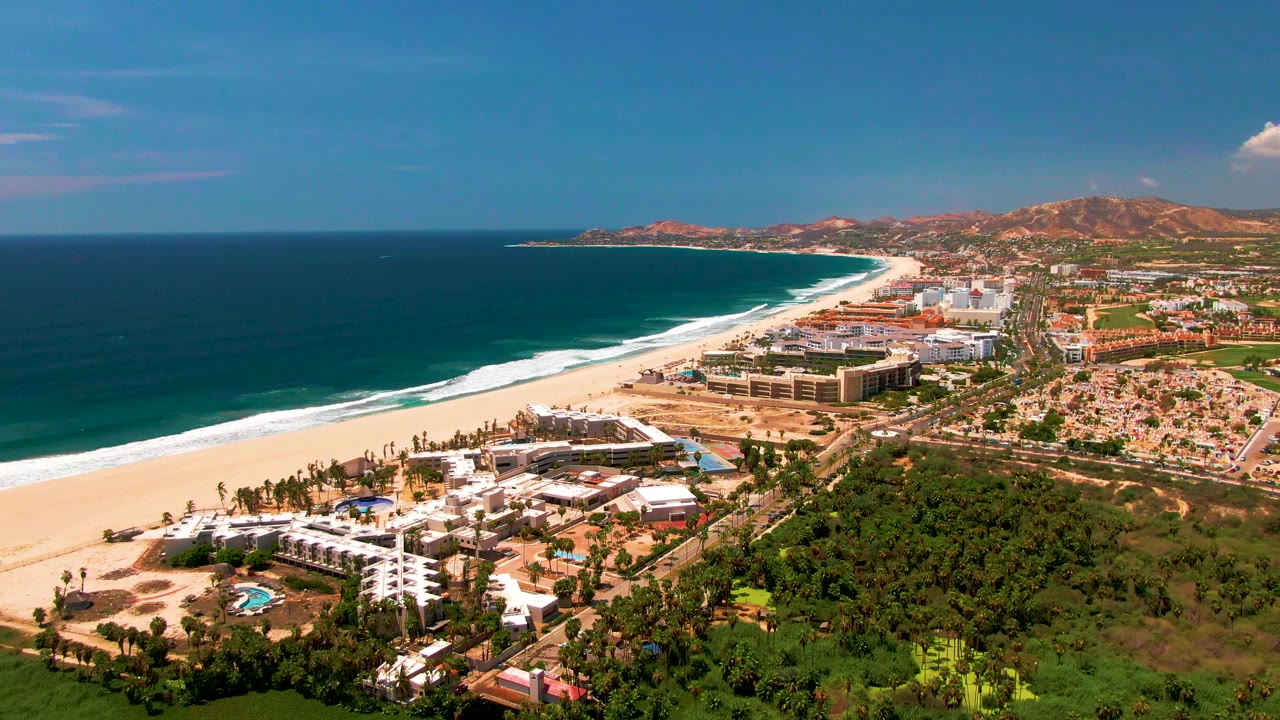 Wide coastal aerial with resort buildings, beach, ocean waves, greenery, and hills.