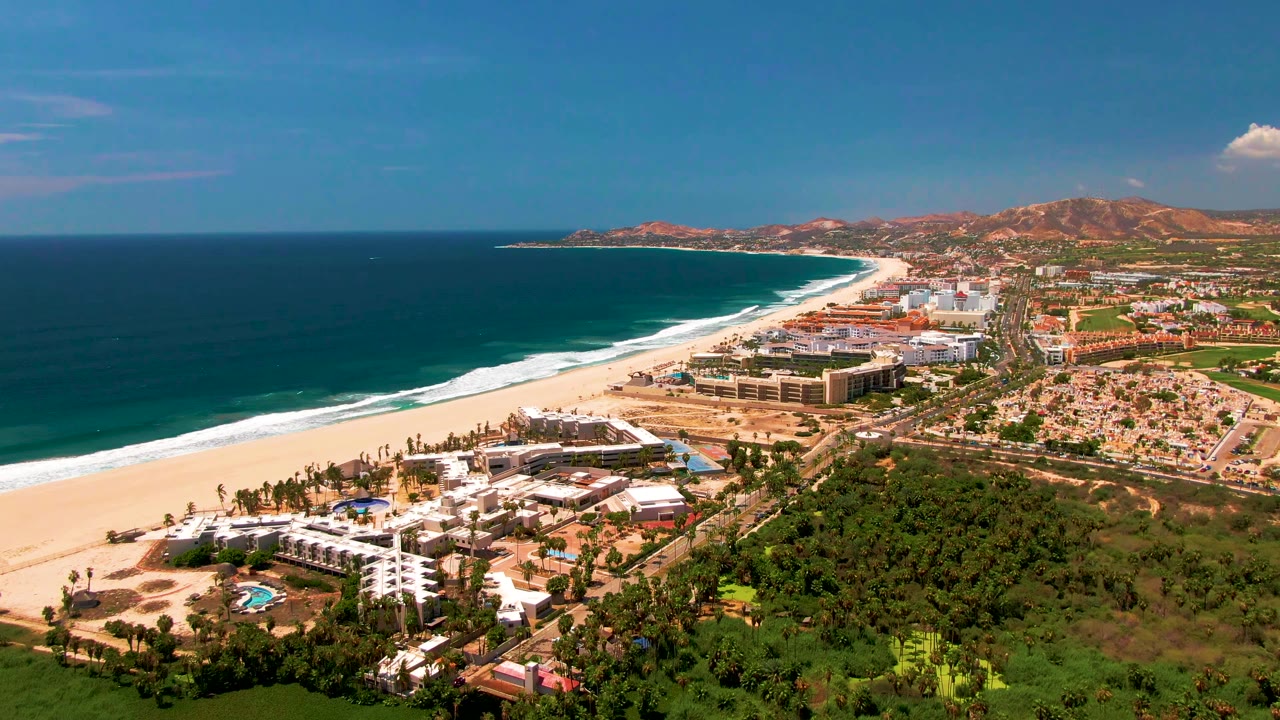 San Jose del Cabo aerial view with dark ocean on the left, bright sand and surf along the shore, resort blocks inland, green vegetation in front, and no visible people.