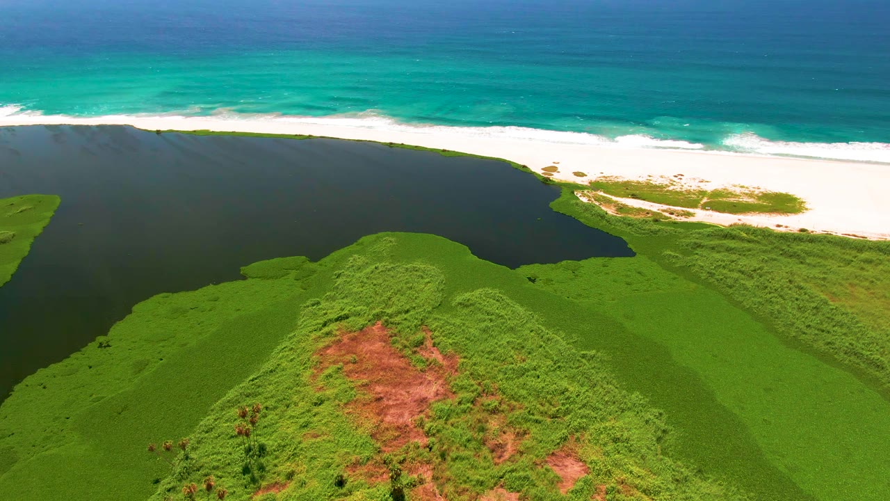 At 0:00, an aerial view in San Jose del Cabo shows green hills in the foreground, dark lagoon water near the beach, turquoise ocean beyond, and no visible people.