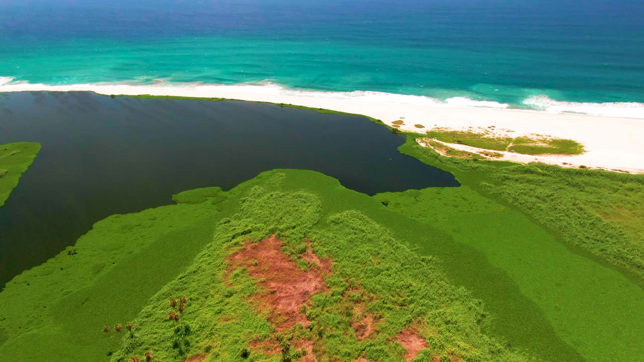 At 0:02, green hills and a reddish earth patch fill the lower frame while the lagoon, white beach, and turquoise ocean stack into horizontal bands in San Jose del Cabo, with no visible people.