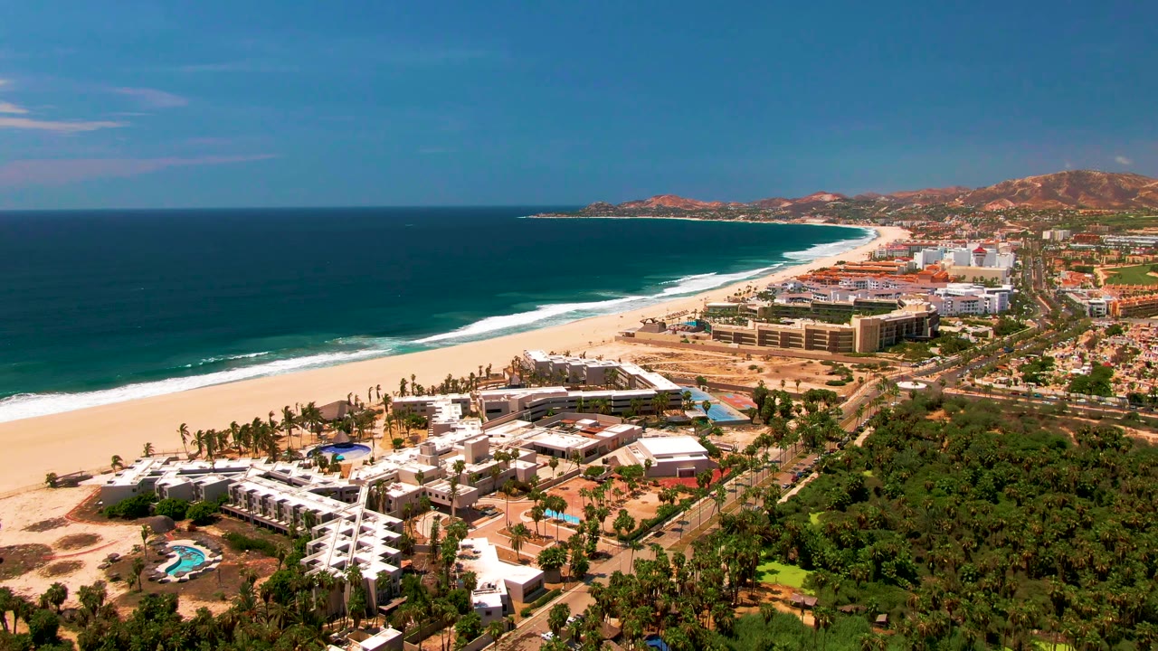 A wide aerial frame over San Jose Del Cabo shows white resort buildings, a pool, green trees, a sandy beach, blue ocean, inland roads, and distant hills with no visible people.