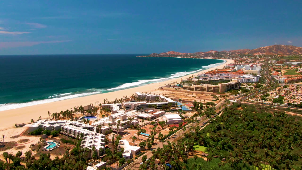 At 0:01, the San Jose Del Cabo aerial view keeps the beach and surf line prominent, with resort buildings and a small pool below, inland roads to the right, and no visible people.