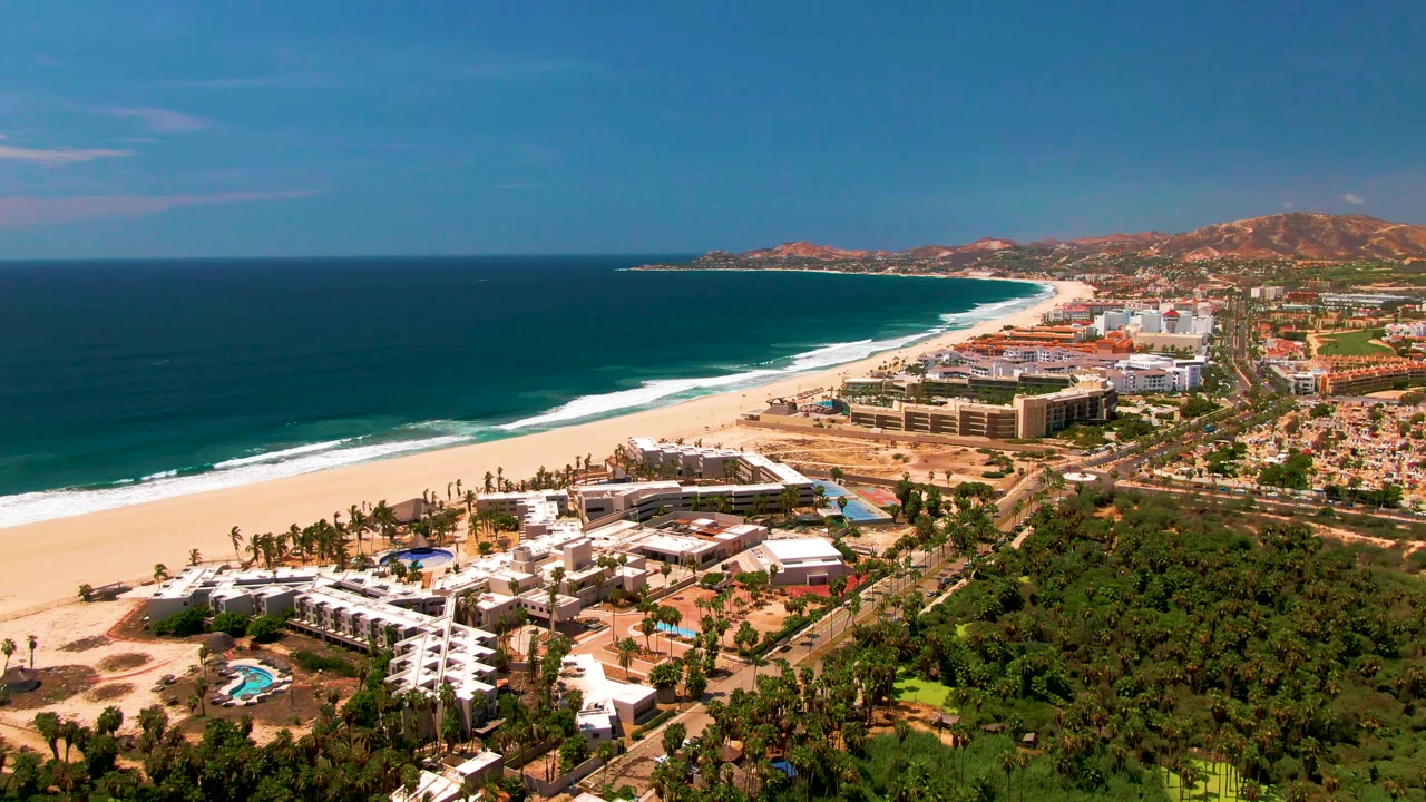 At 0:02, the frame shows San Jose Del Cabo resort blocks near the sand, the ocean stretching left of frame, greenery in the lower right, reddish hills beyond town, and no visible people.