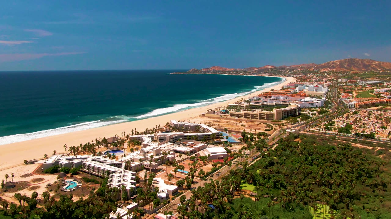 Drone-style view over white resort buildings, pool, beach, ocean, and inland greenery.