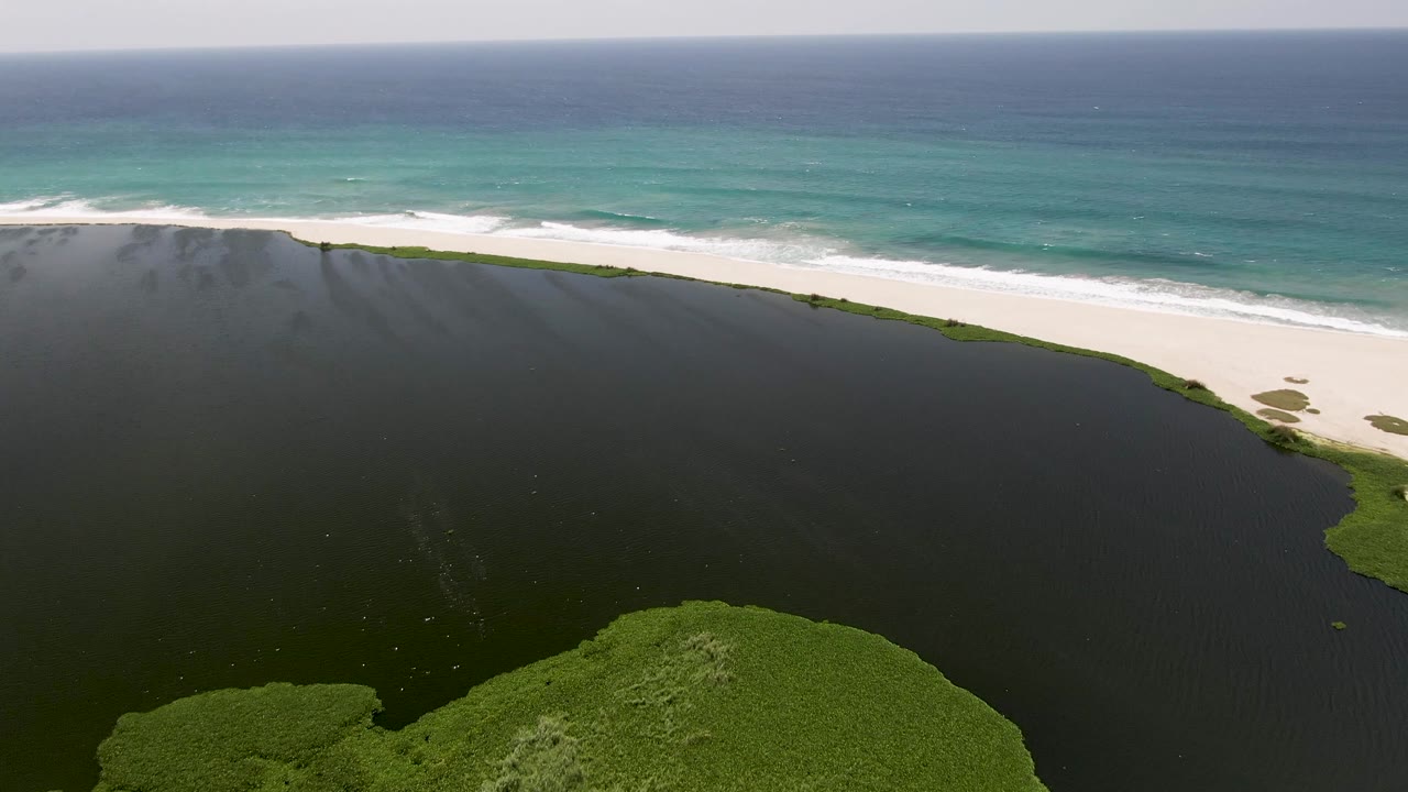 San Jose del Cabo coastline from above, with a broad dark lagoon in the foreground, a thin white sand beach across the frame, blue-green ocean waves, and no visible people.