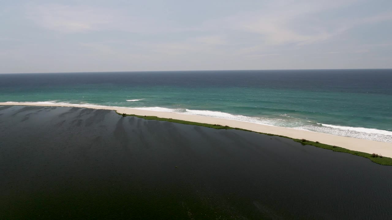 Aerial view of dark cliff edge, sandy beach, and turquoise ocean surf.