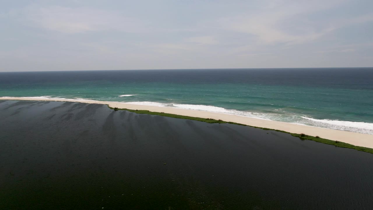 At 0:02, an elevated San Jose del Cabo coastal frame shows dark wet foreground terrain, a narrow sandy beach, breaking blue-green waves, and no visible people.