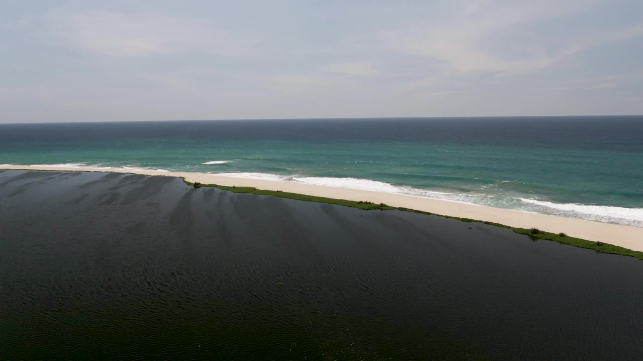 At 0:03, the San Jose del Cabo beach is seen from above with the cliff edge low in frame, wave foam spread farther across the sand, and no visible people.