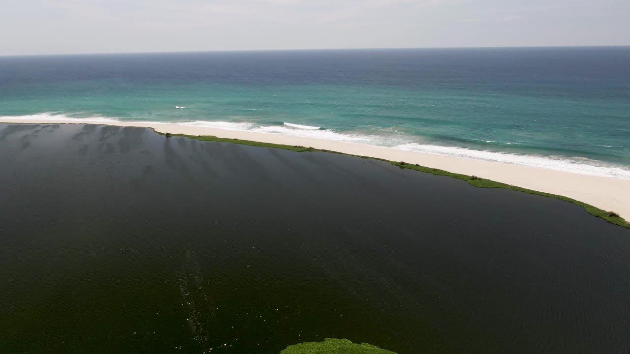 Aerial frame from San Jose del Cabo showing dark lagoon water in the foreground, a white sand beach, turquoise ocean waves, and no visible people.