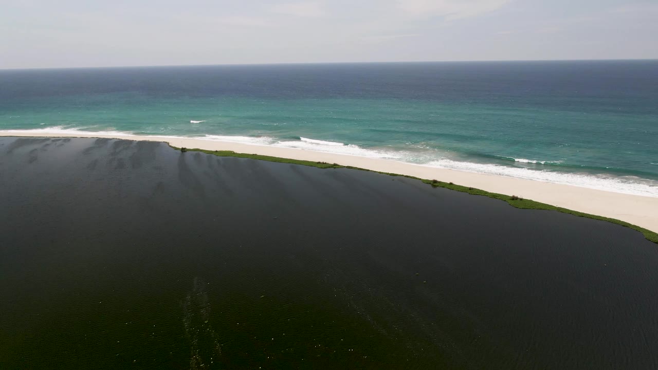 Wide aerial view in San Jose del Cabo with dark lagoon water below, a green shoreline strip, white beach, ocean surf, and no visible people.