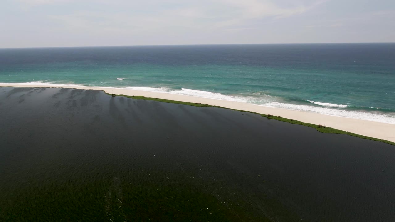 San Jose del Cabo aerial frame showing the dark lagoon foreground, pale beach band, breaking white surf, teal ocean water, and no visible people.