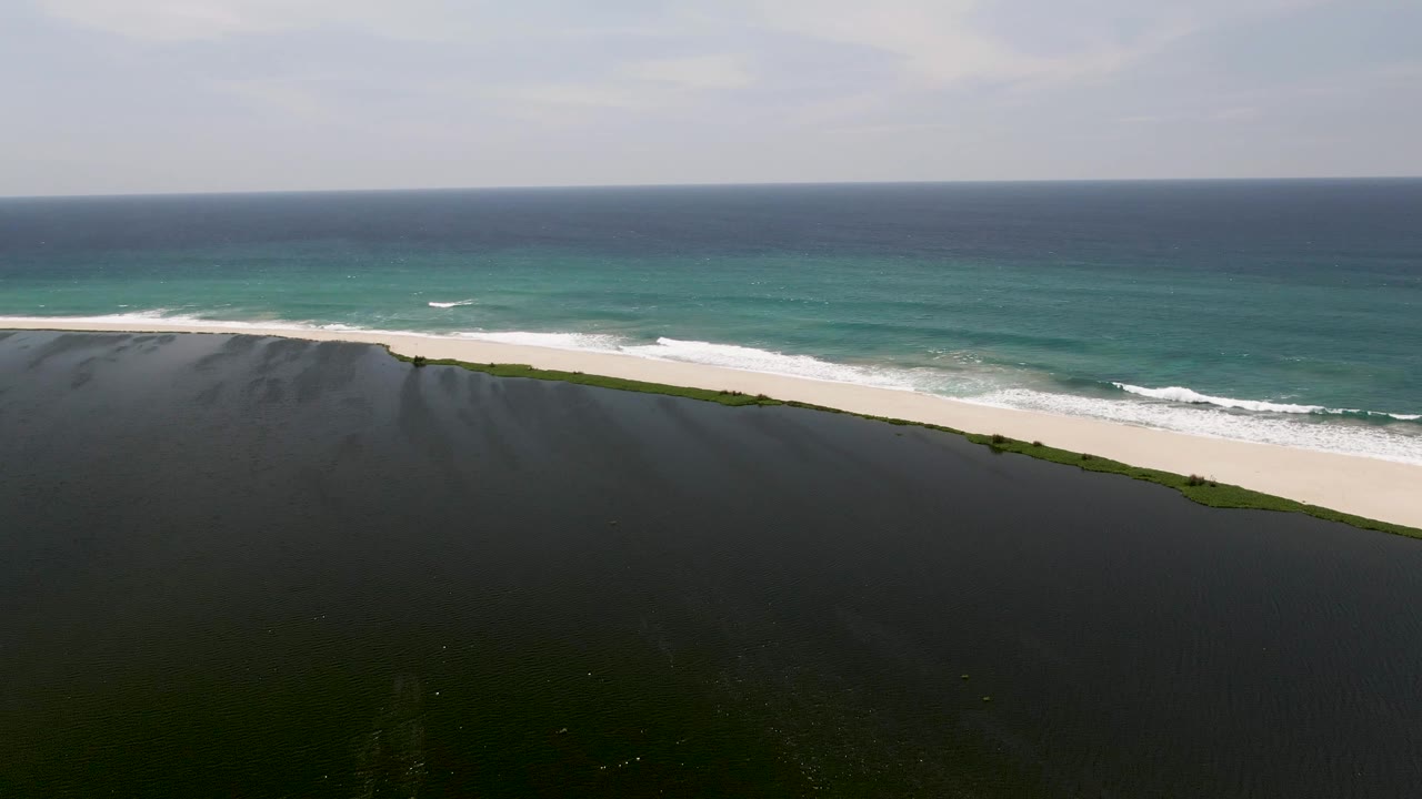 Drone view across lagoon toward white sand and turquoise ocean under cloudy sky.