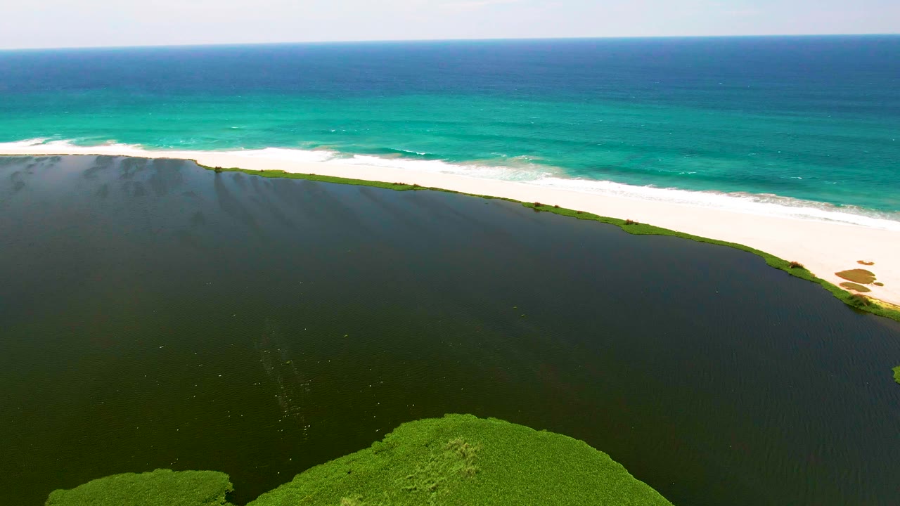 Aerial frame over San Jose Del Cabo showing a dark lagoon in the foreground, a curved white sand beach, turquoise ocean beyond it, and no visible people.