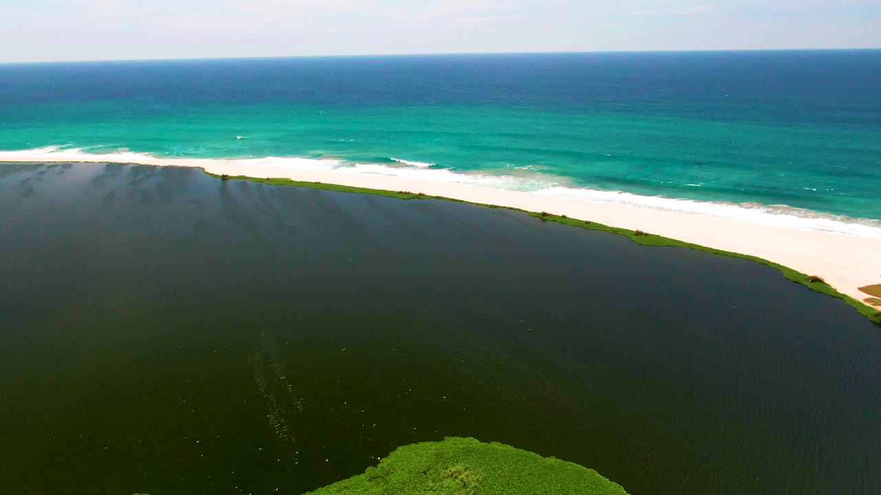 San Jose Del Cabo aerial view with the lagoon filling the lower frame, a narrow green-edged sandbar through the middle, turquoise surf above, and no visible people.