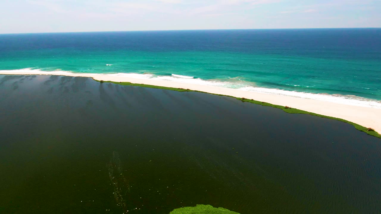 Wide San Jose Del Cabo aerial frame showing dark lagoon water below, a pale beach running diagonally across the scene, ocean waves breaking behind it, and no visible people.