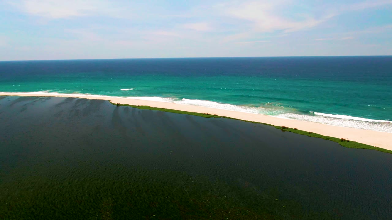 Aerial frame over San Jose del Cabo shoreline with dark water in the foreground, a narrow sandy beach, turquoise surf, blue ocean, thin clouds, and no visible people.