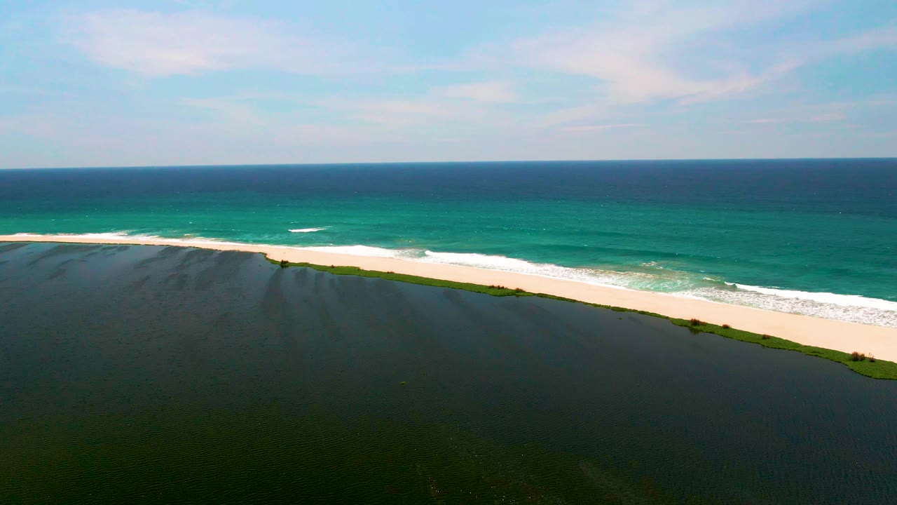 Aerial frame of San Jose del Cabo beach with white foam curling along the pale sand, dark green water below, blue ocean beyond, and no visible people.