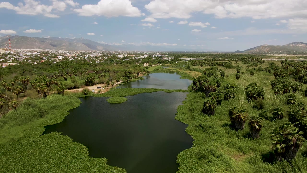 A wide aerial frame over the San Jose del Cabo lake shows dense green banks, palms to the right, a distant city strip, hazy mountains, scattered clouds, and no visible people.