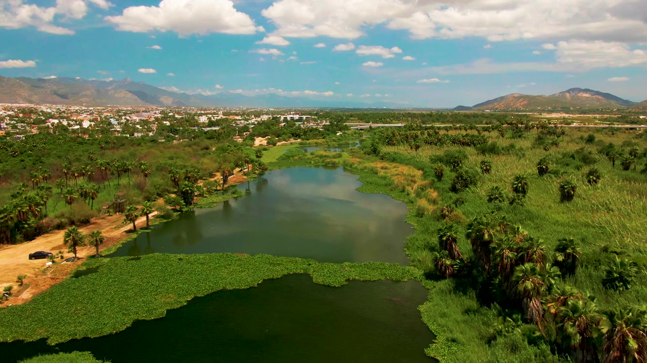 Wide aerial landscape with green water, vegetation, dirt path, town, and mountains.