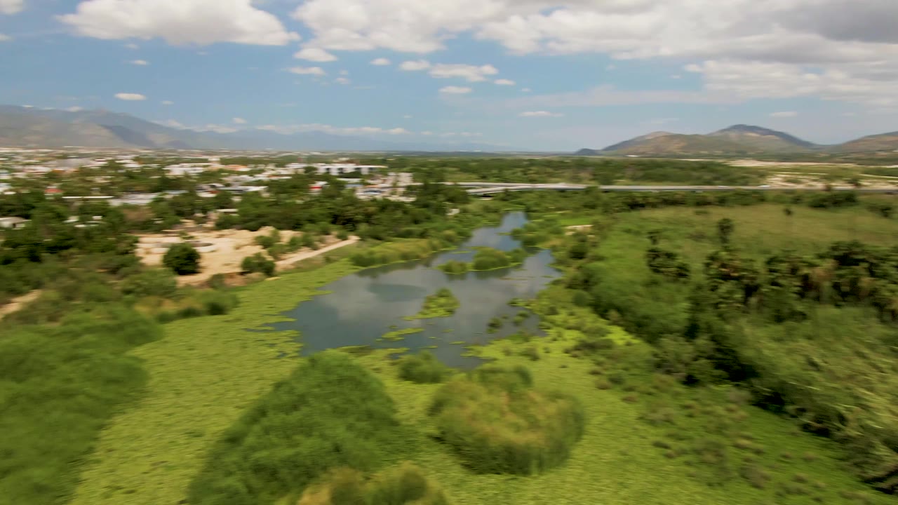 Aerial frame over San Jose Del Cabo showing a small lake at left, dense green vegetation below, distant town buildings, mountains, clouds, and no visible people.
