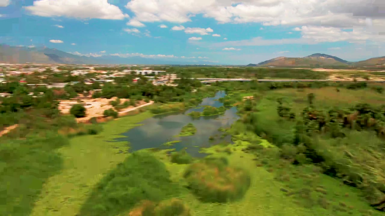 Aerial view over a green pond and wet fields in San Jose del Cabo, with town buildings and hazy mountains beyond under a blue sky, no visible people.
