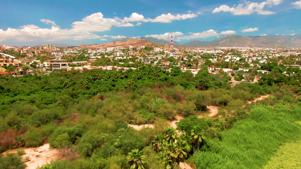 Aerial landscape in San Jose del Cabo with dense green vegetation in front, town buildings and a red-and-white tower in the midground, and mountains under scattered clouds, no visible people.
