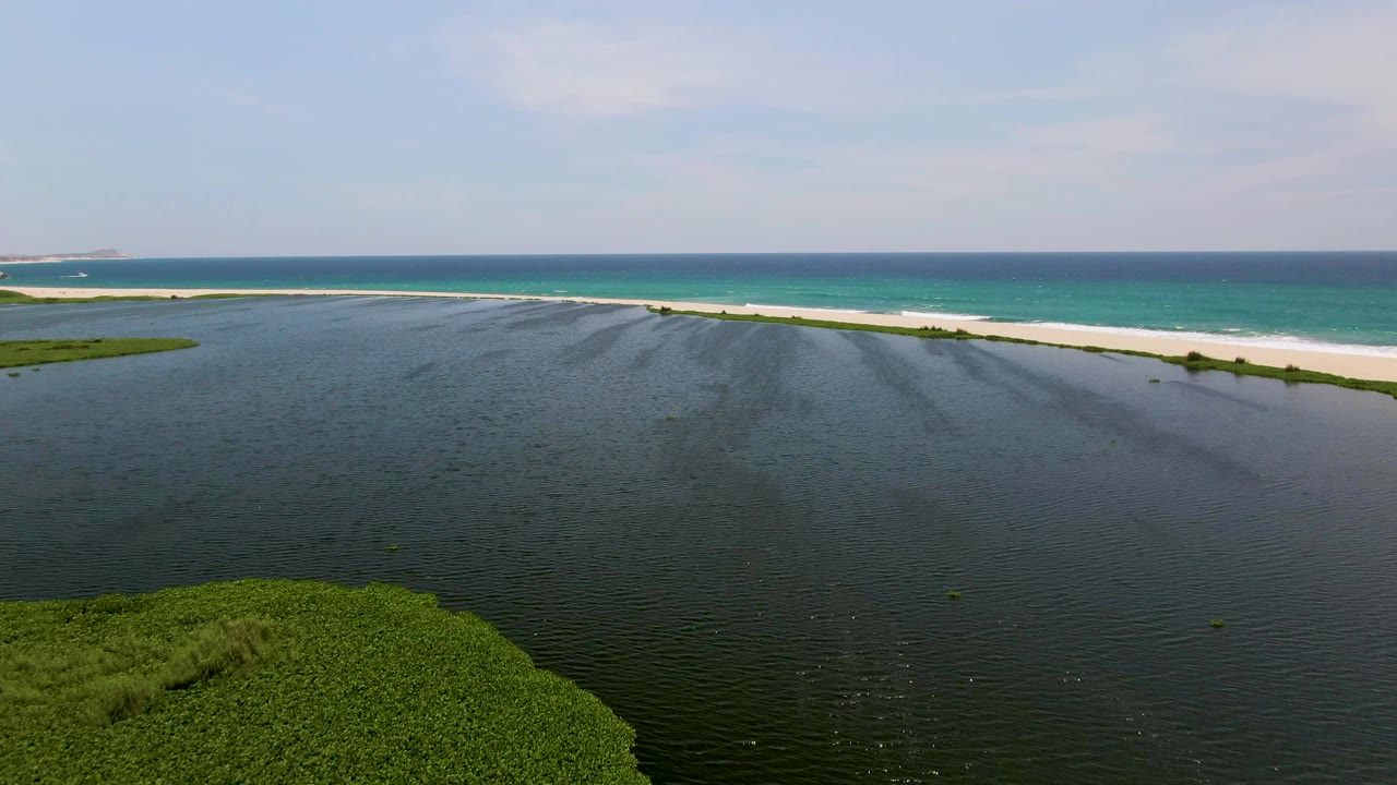 Aerial view in San Jose Del Cabo showing dark rippled lagoon water in the foreground, green vegetation at lower left, a pale sand strip, turquoise ocean beyond, and no visible people.