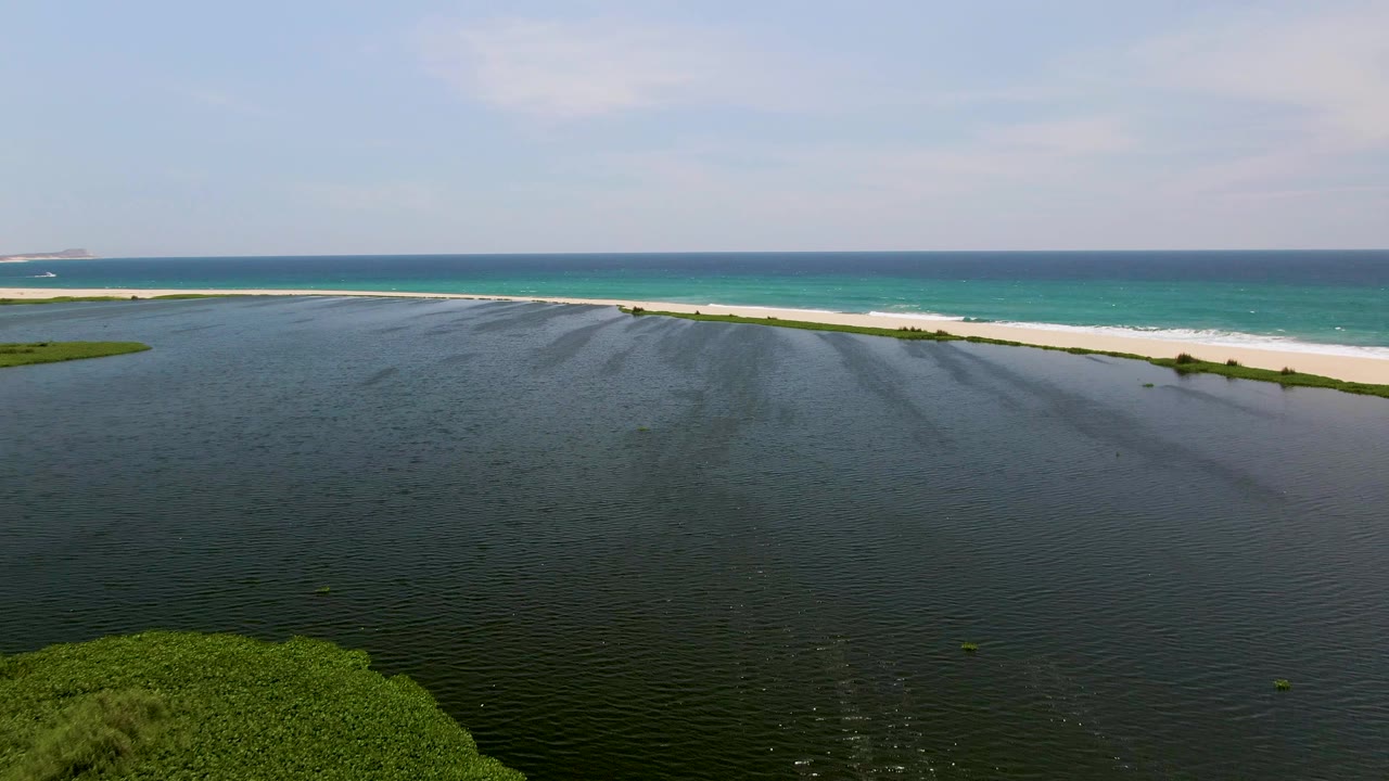 Lagoon water and green shoreline vegetation below a narrow sandbar and blue ocean.
