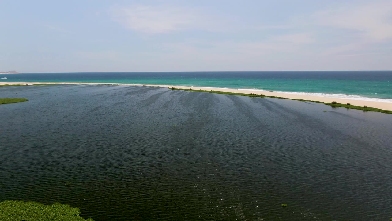 San Jose Del Cabo aerial frame showing a broad dark lagoon surface, small green patches near the shore, a bright sand strip, a thin surf line, turquoise ocean, and no visible people.