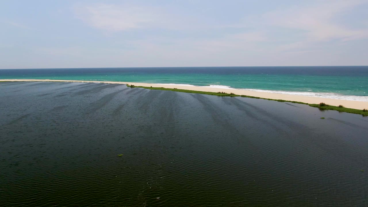 San Jose del Cabo aerial view with rippled dark water in the foreground, a narrow sandy shore with small green patches, turquoise ocean, and no visible people.