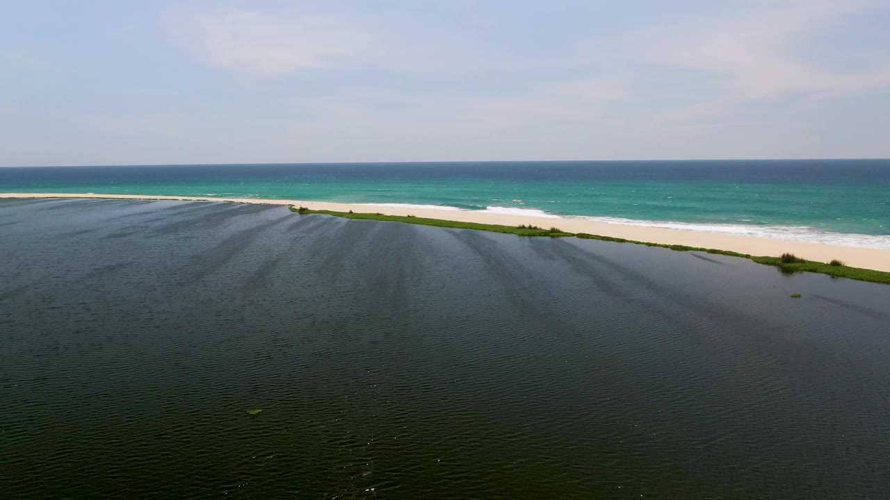 Aerial San Jose del Cabo frame showing the dark shallow water surface, pale sandbar, low green vegetation, bright turquoise sea, and no visible people.