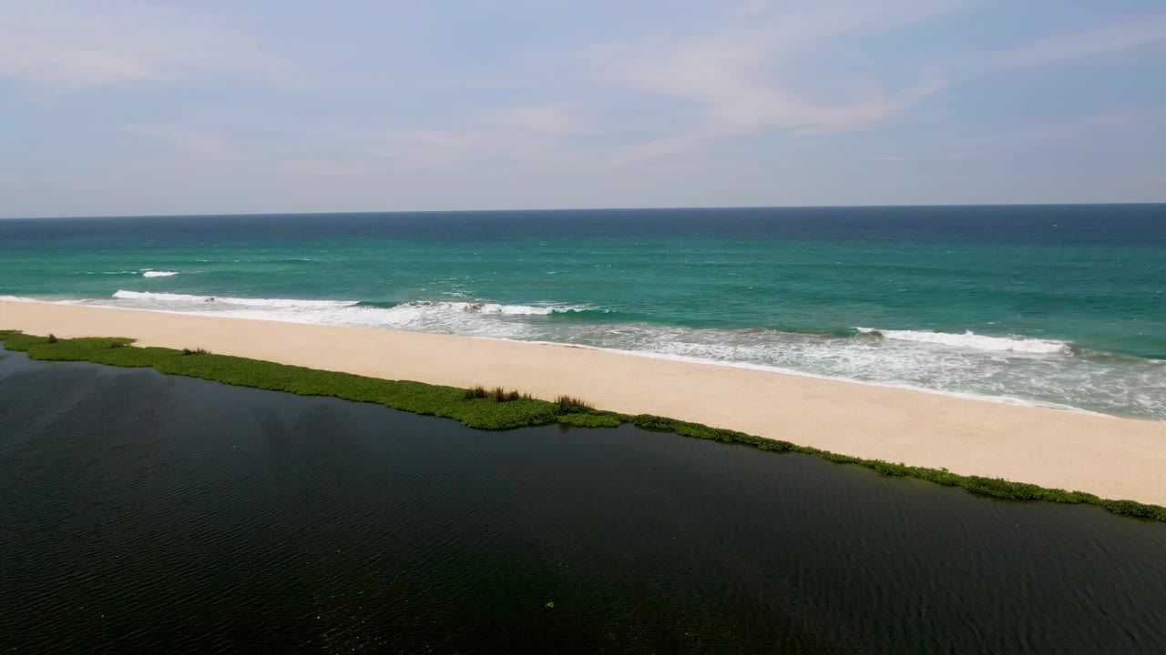 Aerial view in San Jose del Cabo with the lagoon filling the lower frame, a strip of green vegetation, pale sand, white wave foam, and no visible people.