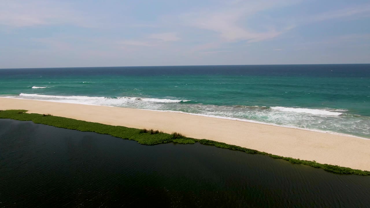 San Jose del Cabo coastline from above, with dark rippled lagoon water at the bottom, green shoreline growth, broad sand, turquoise sea, and no visible people.