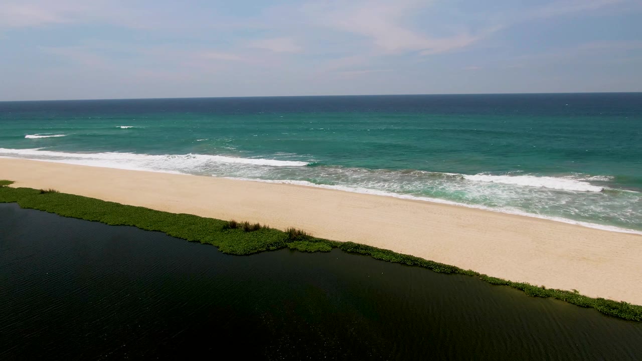 Aerial San Jose del Cabo beach frame with more open surf visible across the upper half, lagoon and vegetation below, pale sand between them, and no visible people.
