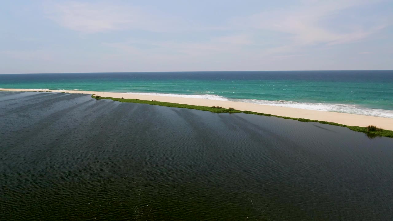 Aerial frame from San Jose Del Cabo showing dark lagoon water in the foreground, a pale sand beach, turquoise ocean beyond, and no visible people.