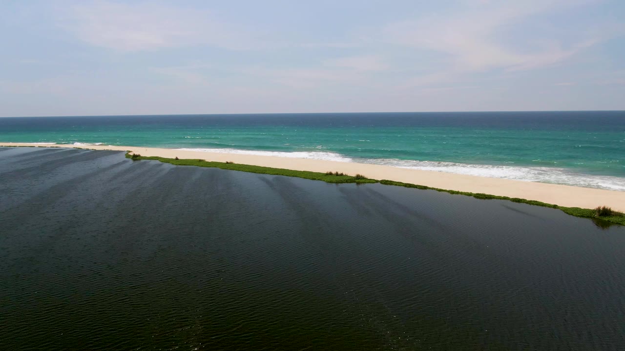 Aerial view in San Jose Del Cabo with rippled lagoon water below, green vegetation along the shore, bright sand, rolling surf, and no visible people.