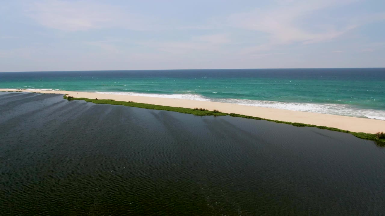 San Jose Del Cabo aerial frame with the lagoon occupying the lower half, a thin sand strip across the middle, ocean waves breaking behind it, and no visible people.