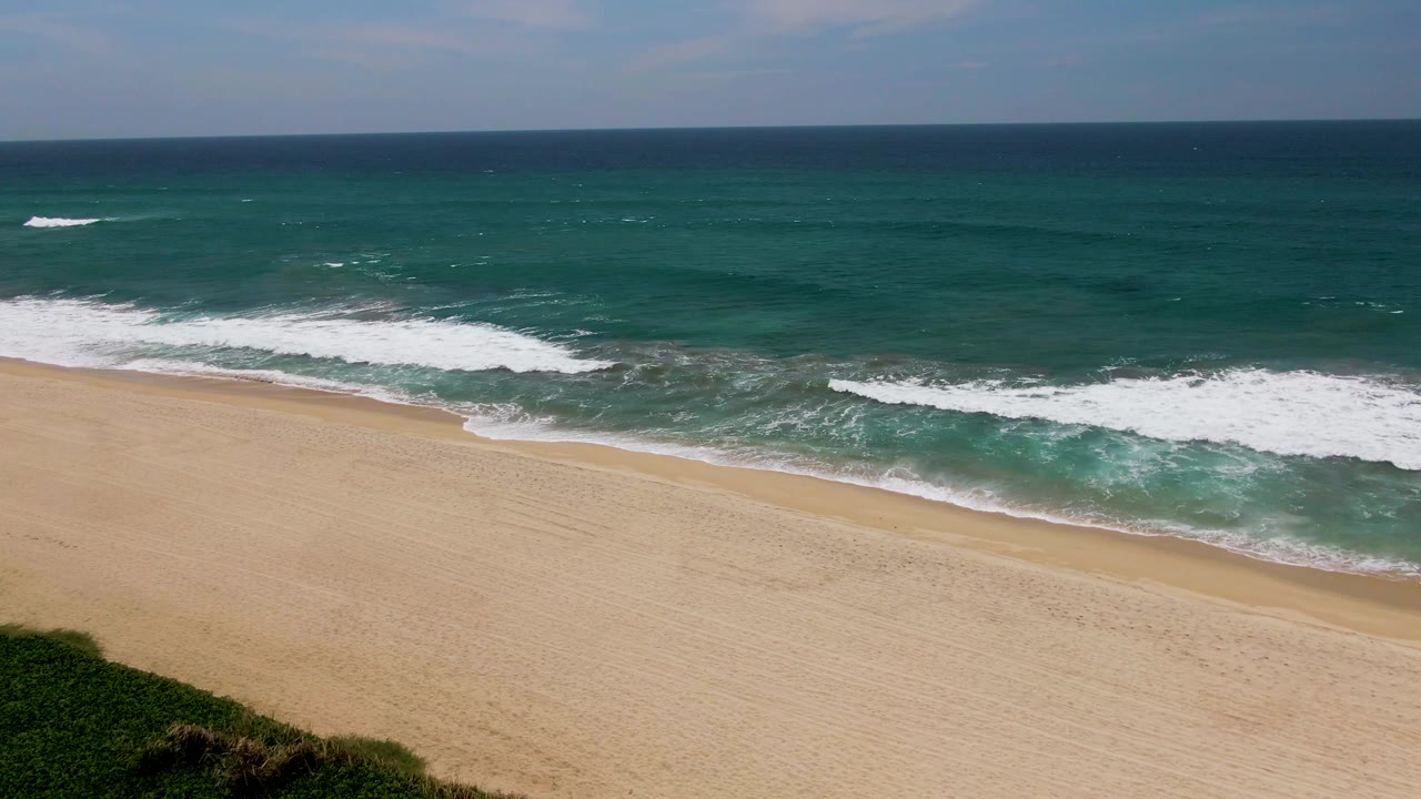 White foam spreads across turquoise water near a wide sandy beach in San Jose Del Cabo, with deep ocean beyond and no visible people.