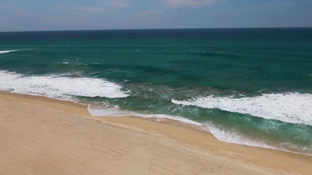 Breaking surf moves closer to the empty sandy shoreline in San Jose Del Cabo, with white foam filling more of the lower ocean area and no visible people.