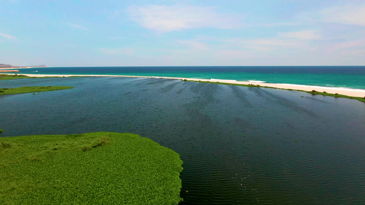 Aerial frame over San Jose del Cabo lagoon water with a green vegetated mass in the lower left, a white sandbar across the middle, turquoise ocean beyond, and no visible people.