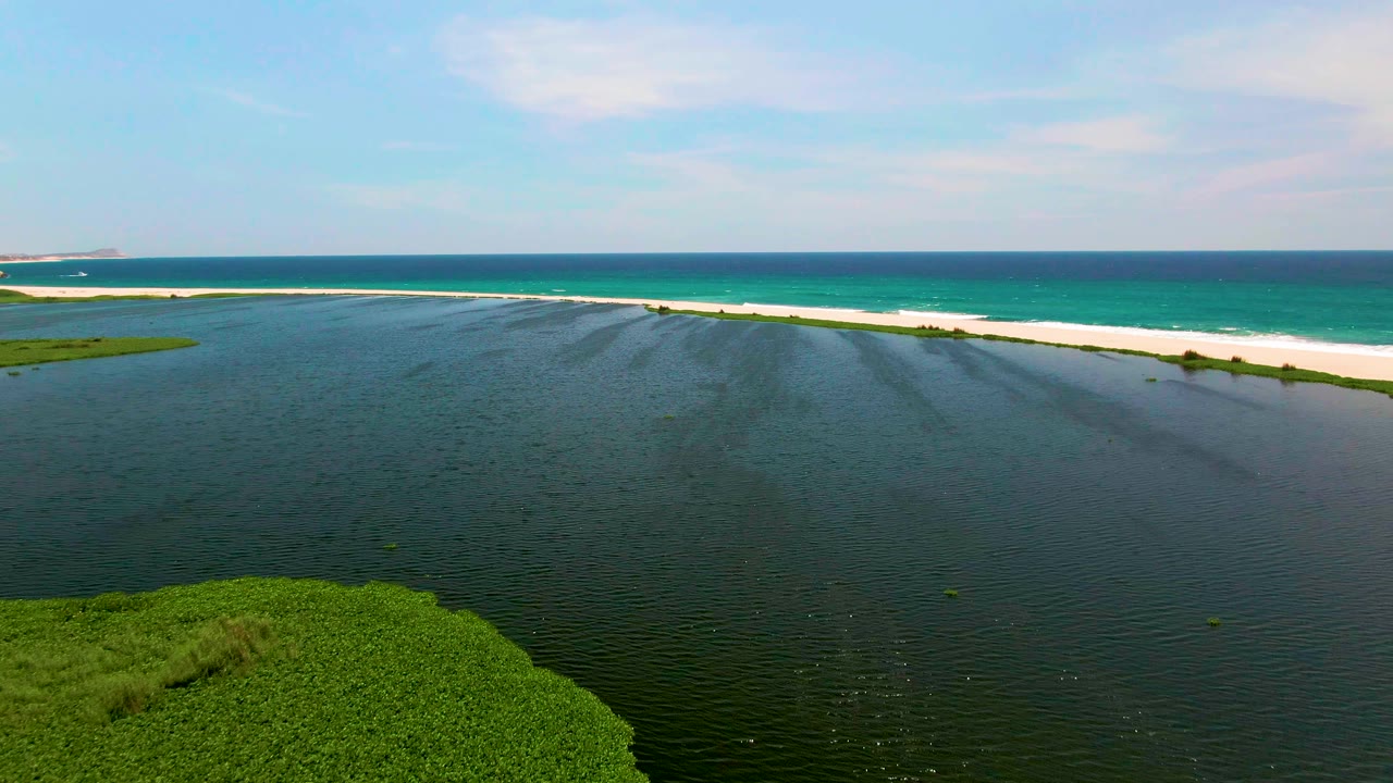 San Jose del Cabo aerial frame showing broad rippled lagoon water, a narrow white sandbar, blue-green ocean, a small green shoreline strip at left, and no visible people.
