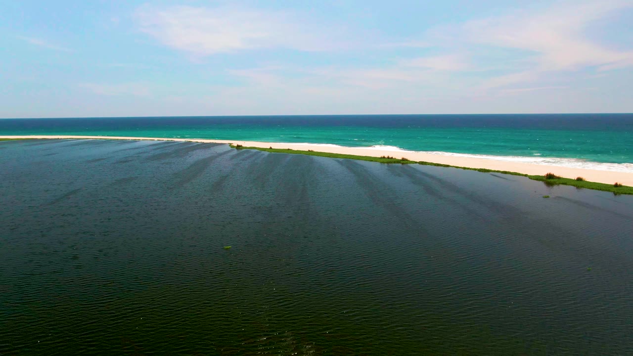 Aerial frame from San Jose del Cabo showing dark green lagoon water in front, a pale sand beach with low vegetation, turquoise ocean beyond, and no visible people.