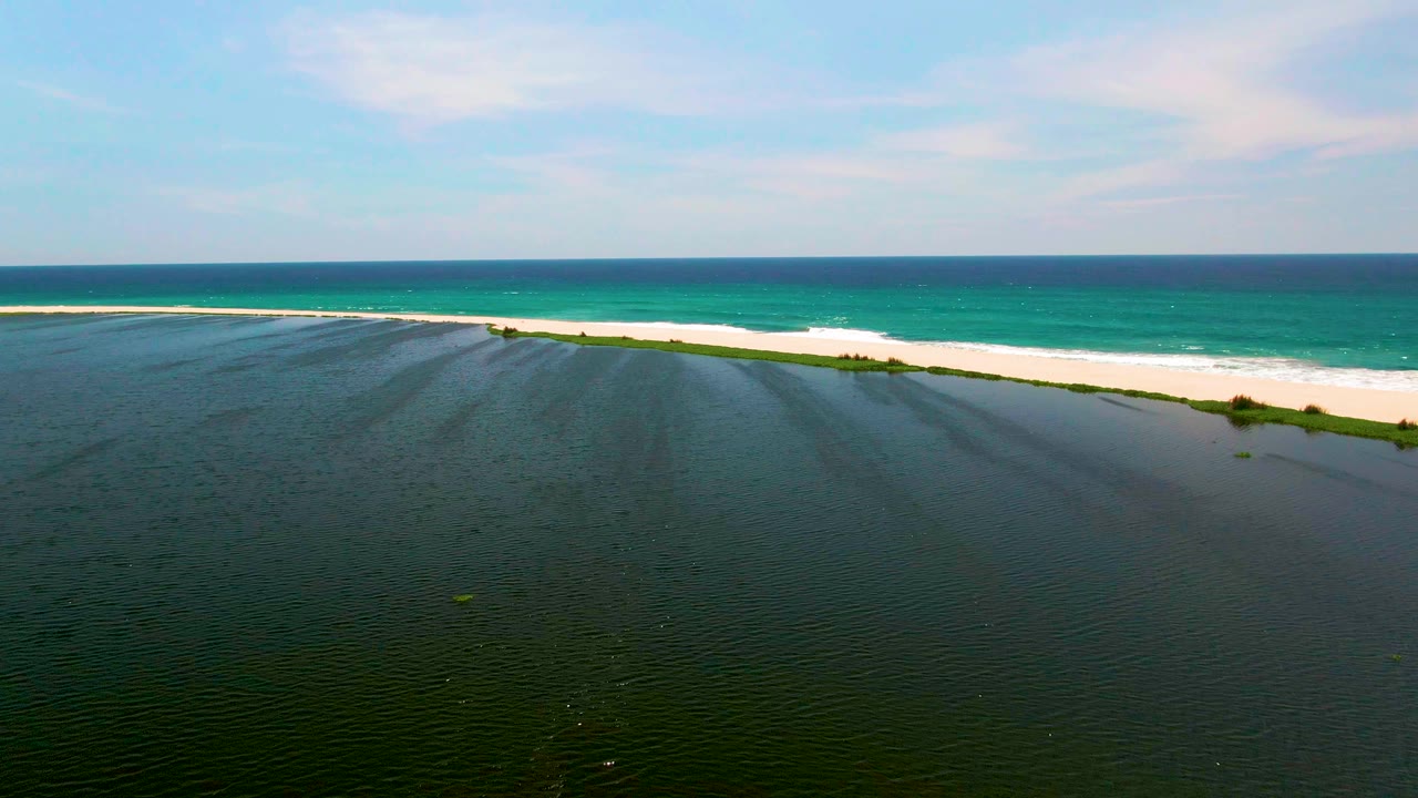 A wide San Jose del Cabo aerial still holds the lagoon across the lower frame, the sandbar through the center, a white surf line at the beach, and no visible people.