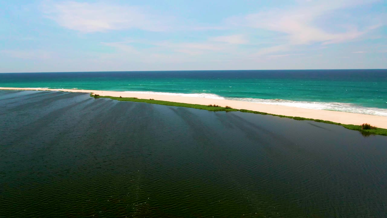 An aerial frame from San Jose del Cabo shows dark rippled lagoon water in front of a pale sand strip and turquoise ocean waves beyond, with no visible people.