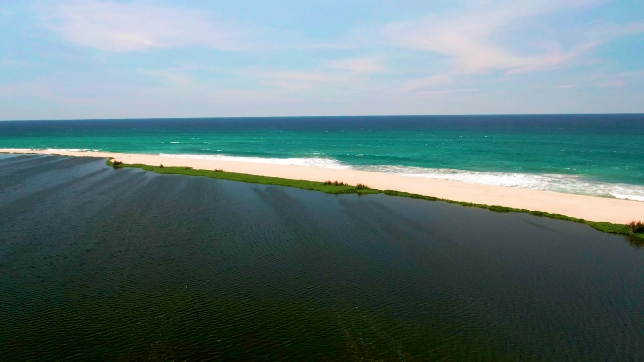 At 0:02, the aerial view shows more of the diagonal beach edge between rippled lagoon water and teal ocean waves under a lightly clouded sky, with no visible people.