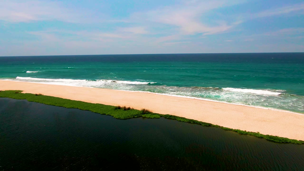 A high aerial frame in San Jose del Cabo shows dark lagoon water, green shoreline vegetation, a broad sandy beach, turquoise ocean surf, blue sky, and no visible people.