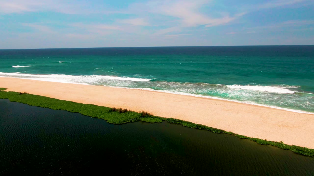 Aerial footage over San Jose del Cabo frames the green lagoon edge in the foreground, pale sand across the center, white wave breaks offshore, and no visible people.